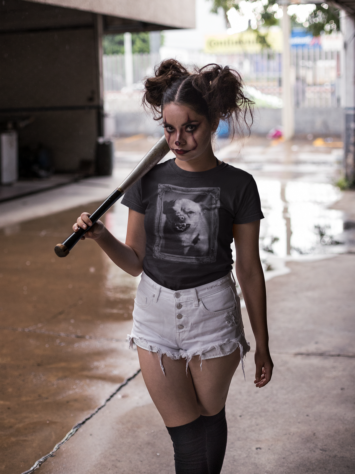 A woman in a zombie-style makeup look stands on a wet urban sidewalk, holding a baseball bat over her shoulder. She is wearing a dark gray graphic t-shirt, white denim shorts, and black knee-high socks.