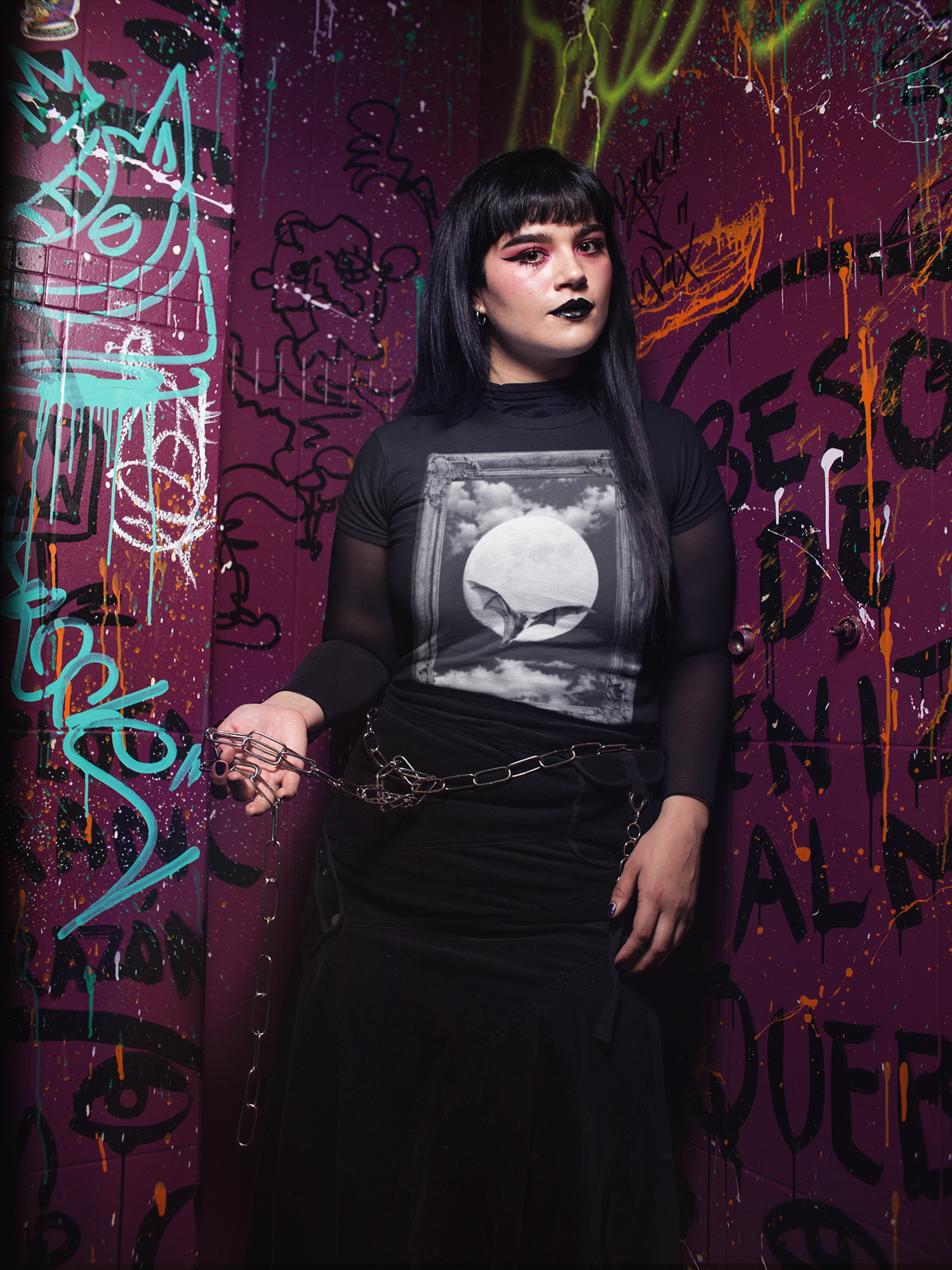 A person with long black hair and black lipstick stands against a graffiti-covered purple wall, wearing a black sheer-sleeve top with a black-and-white graphic print on the front, a black skirt, and a metal chain belt.