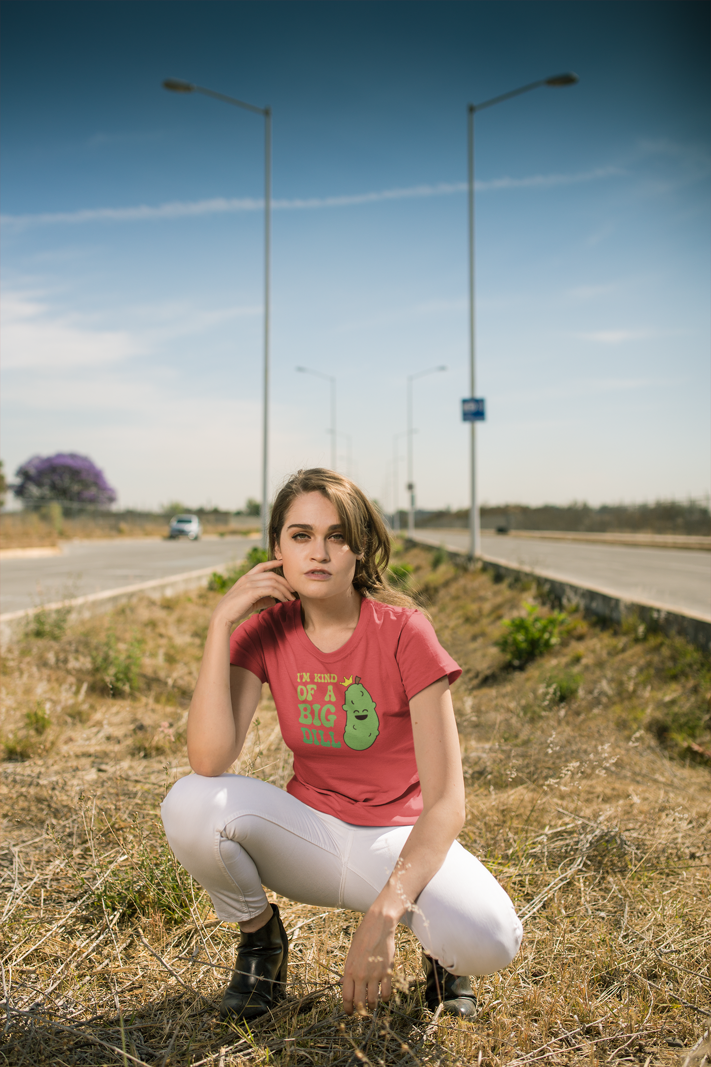 A woman crouching outdoors on a roadside grassy area, wearing a pink graphic t-shirt with a green cartoon dill pickle character and the text 'I'M KIND OF A BIG DILL', paired with white pants and black boots.