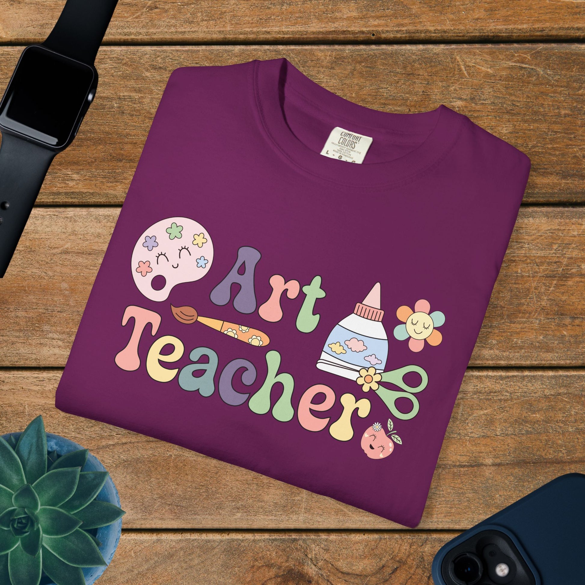 Purple t-shirt with 'Art Teacher' design on a wooden surface with a watch, phone, and plant.