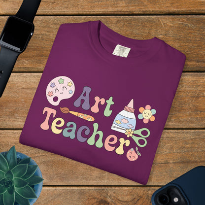 Purple t-shirt with 'Art Teacher' design on a wooden surface with a watch, phone, and plant.