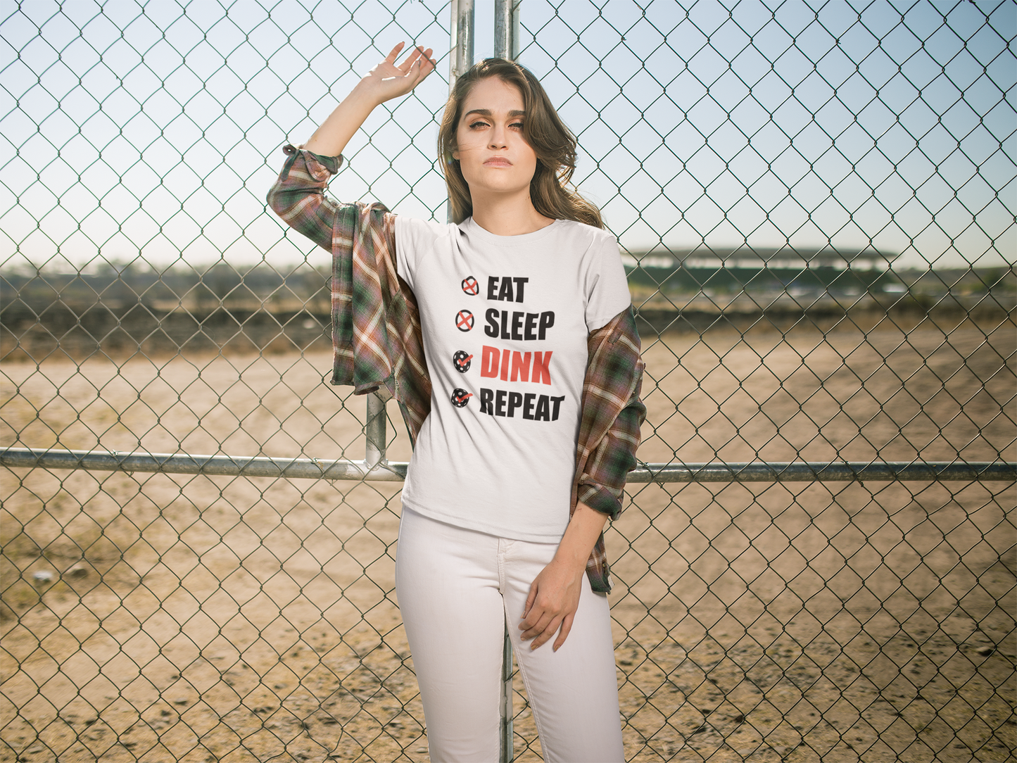 A woman stands in front of a chain-link fence wearing a white graphic t-shirt with the text 'EAT SLEEP DINK REPEAT' in black and red, styled with a plaid shirt draped off her shoulders and white pants.