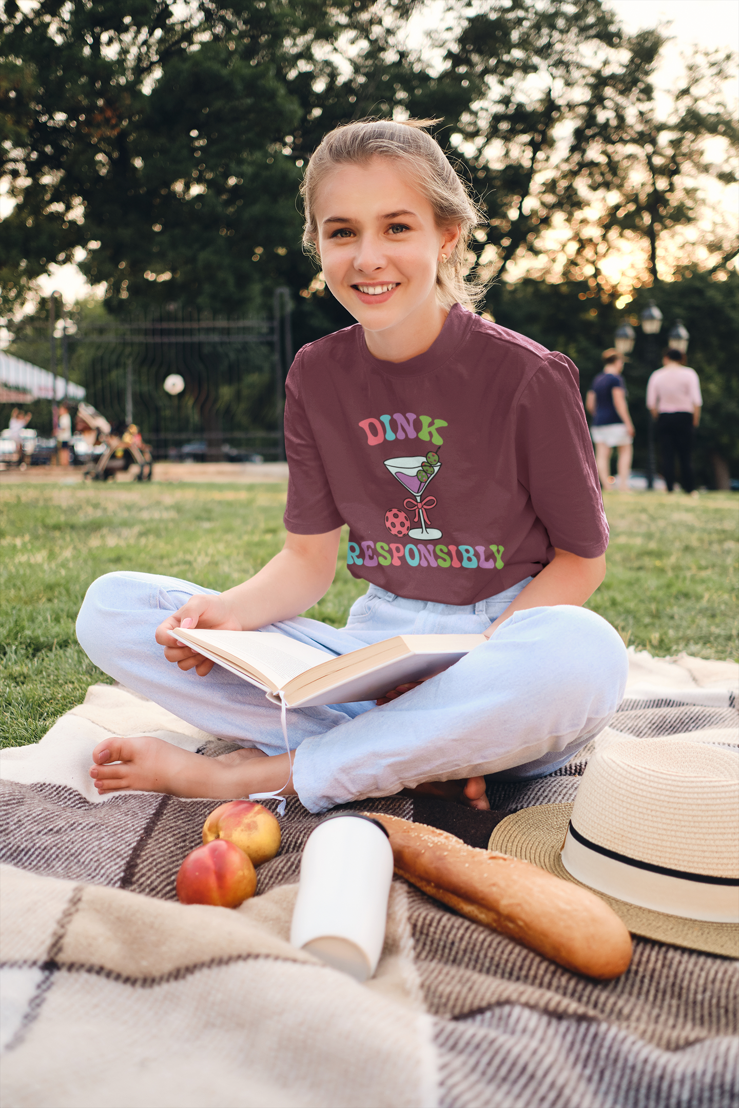 A young woman sits cross-legged on a picnic blanket in a park, wearing a maroon graphic t-shirt that reads 'DINK RESPONSIBLY' with a martini glass illustration. She is holding an open book, with a straw hat, bread, and peaches nearby on the blanket.