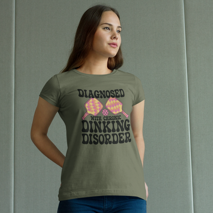 A woman wearing a muted olive green graphic t-shirt with the text 'DIAGNOSED WITH CHRONIC DINKING DISORDER' and a pink and yellow patterned graphic, standing against a gray paneled wall.