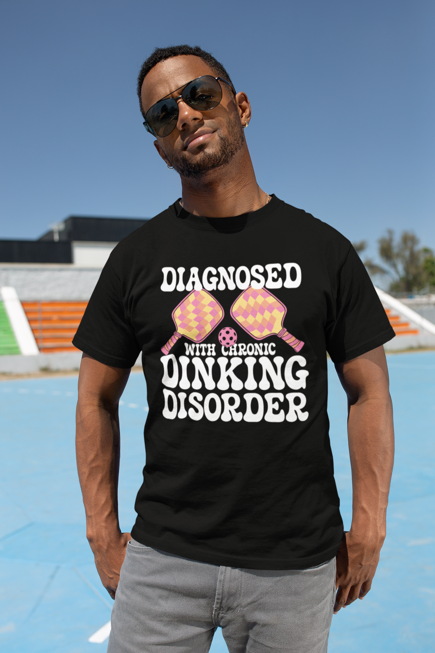 A man wearing a black graphic t-shirt with the text 'DIAGNOSED WITH CHRONIC DINKING DISORDER' and two pink and yellow pince-nez glasses, standing on an outdoor court under a blue sky.