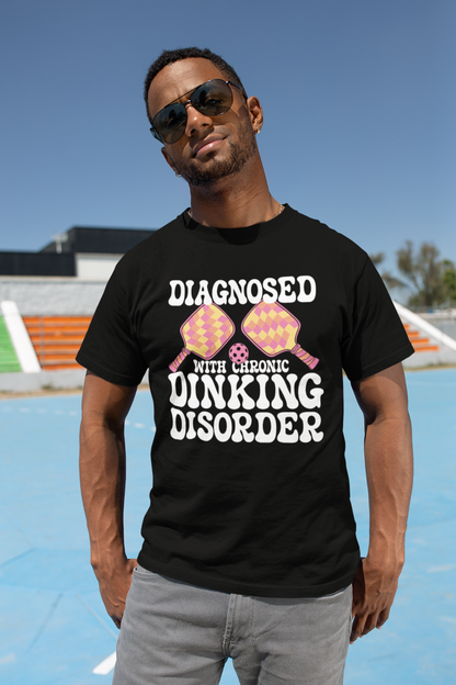 A man wearing a black graphic t-shirt with the text 'DIAGNOSED WITH CHRONIC DINKING DISORDER' and two pink and yellow pince-nez glasses, standing on an outdoor court under a blue sky.