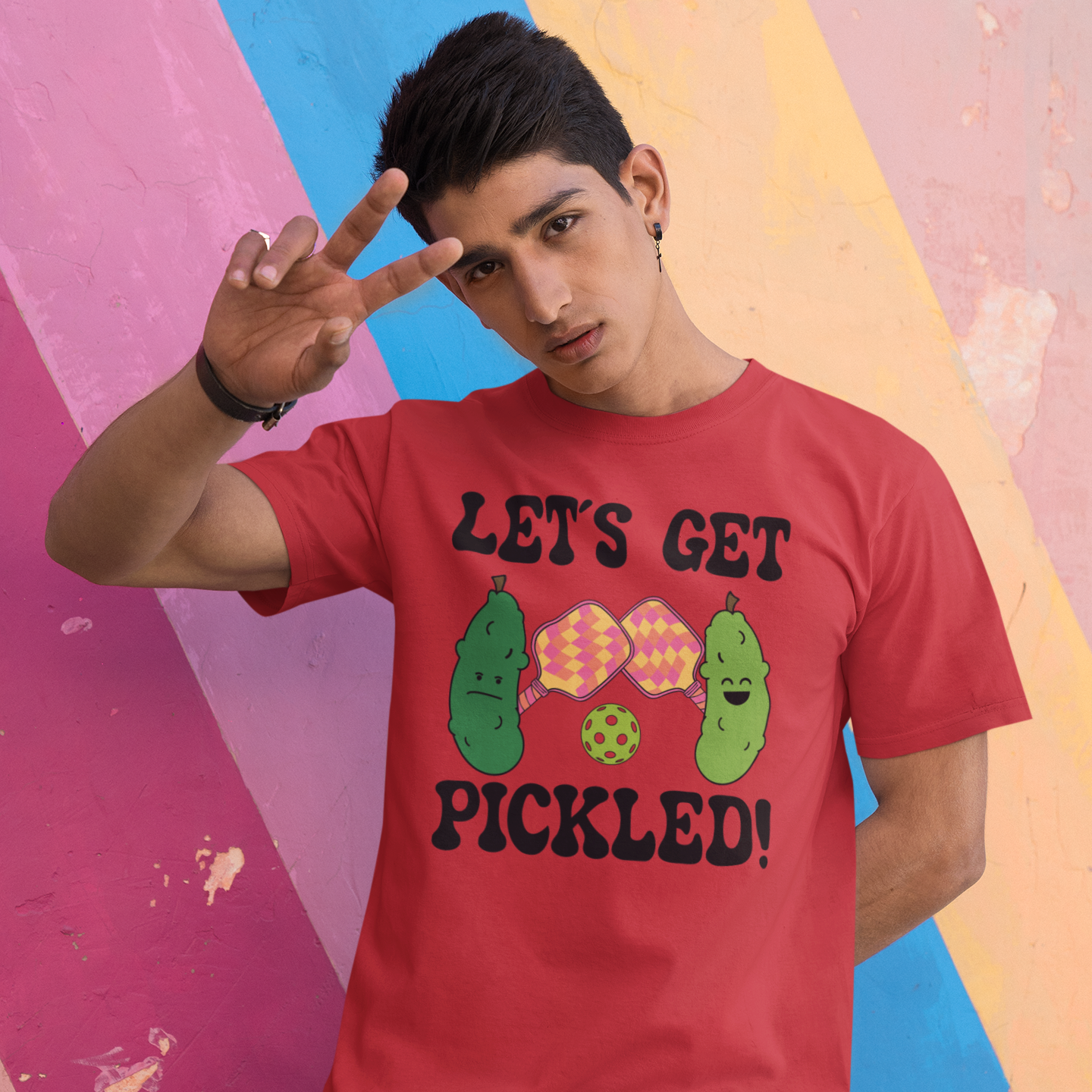 A man wearing a red graphic t-shirt with a cartoon pickle design and the text 'LET'S GET PICKLED!' poses in front of a colorful painted wall.