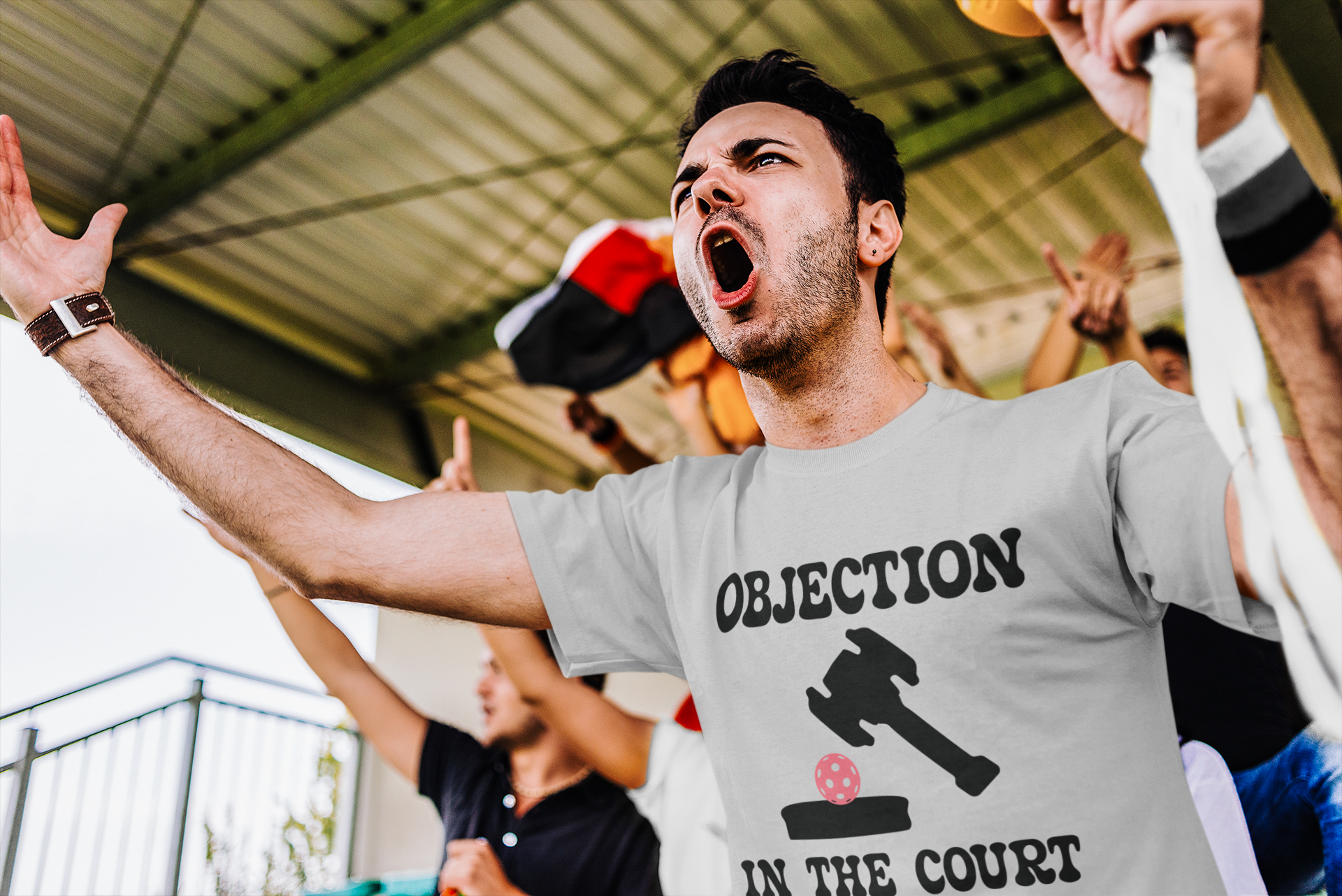 A man in a crowd wearing a light gray t-shirt with 'OBJECTION IN THE COURT' and a gavel graphic printed on the front, shouting with his arms raised.