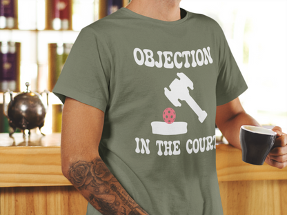 A man wearing an olive green t-shirt with a white graphic that reads 'OBJECTION IN THE COURT' alongside a judge's gavel and gavel ball illustration, holding a black coffee mug.