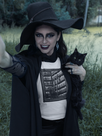 A woman in a witch costume with a wide-brimmed black hat, black cloak, and a graphic t-shirt holds a black cat in an outdoor grassy field. The t-shirt features a black-and-white graphic of a library bookshelf.
