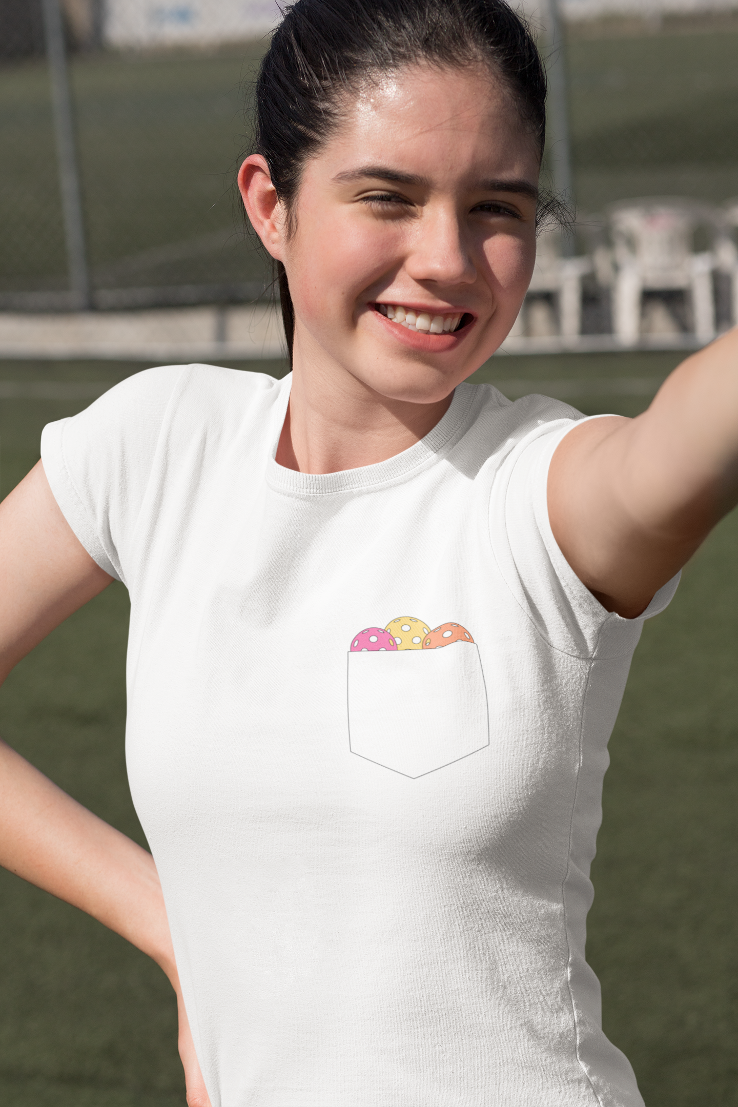 A woman wearing a white short-sleeve t-shirt with a chest pocket containing a graphic of colorful pickleball balls, photographed outdoors on a grassy field.