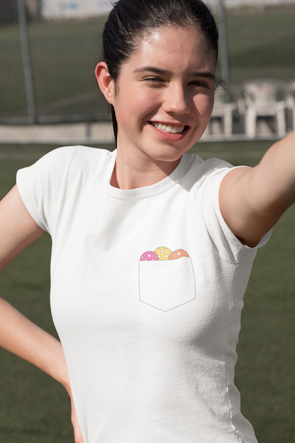 A woman wearing a white short-sleeve t-shirt with a chest pocket containing a graphic of colorful pickleball balls, photographed outdoors on a grassy field.