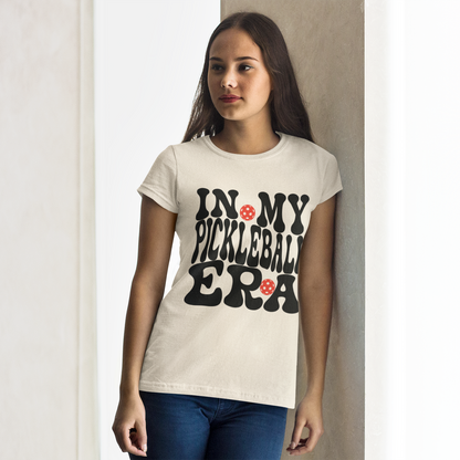 A woman wearing a cream-colored t-shirt with a black graphic text that reads 'IN MY PICKLEBALL ERA' accented with small red flowers, standing against a light-colored wall.