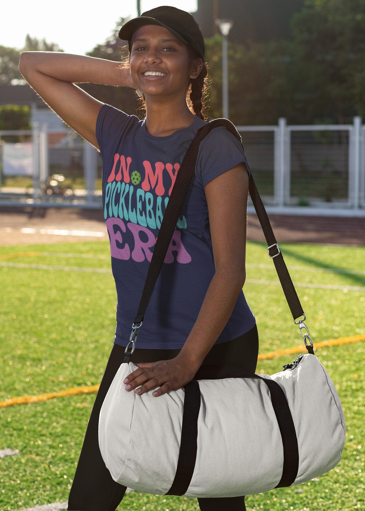 A woman on a sports field carrying a large white duffel bag with black straps and trim. She is wearing a navy graphic t-shirt and a black cap.