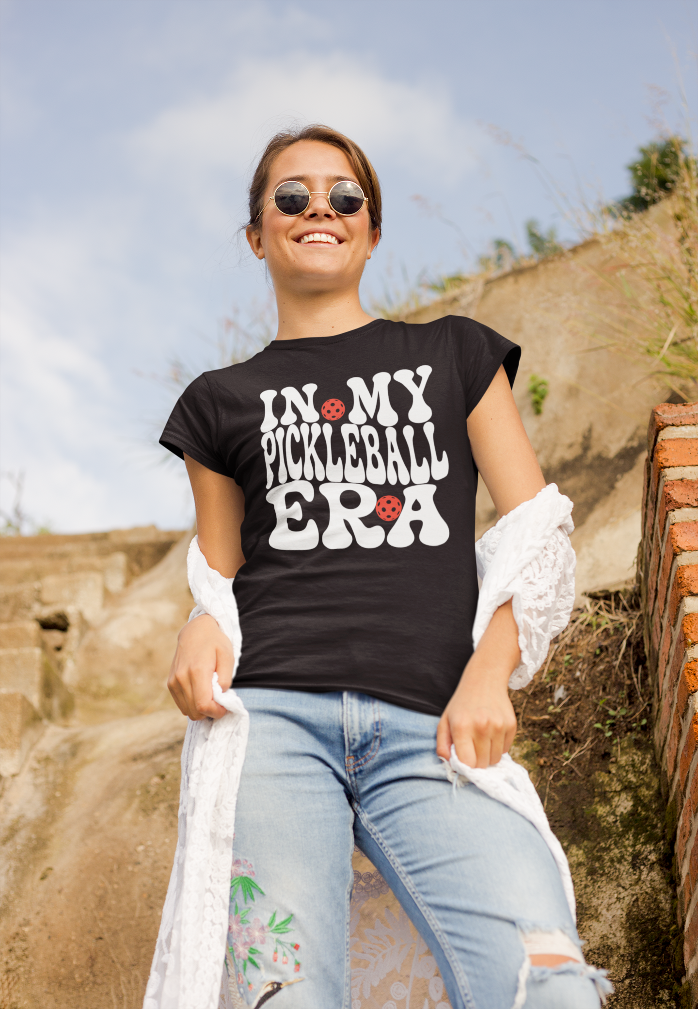 A woman wearing a black graphic t-shirt with the text 'IN MY PICKLEBALL ERA' in white and red, paired with light blue jeans and a white lace shirt draped off her shoulders, set against an outdoor rocky background.