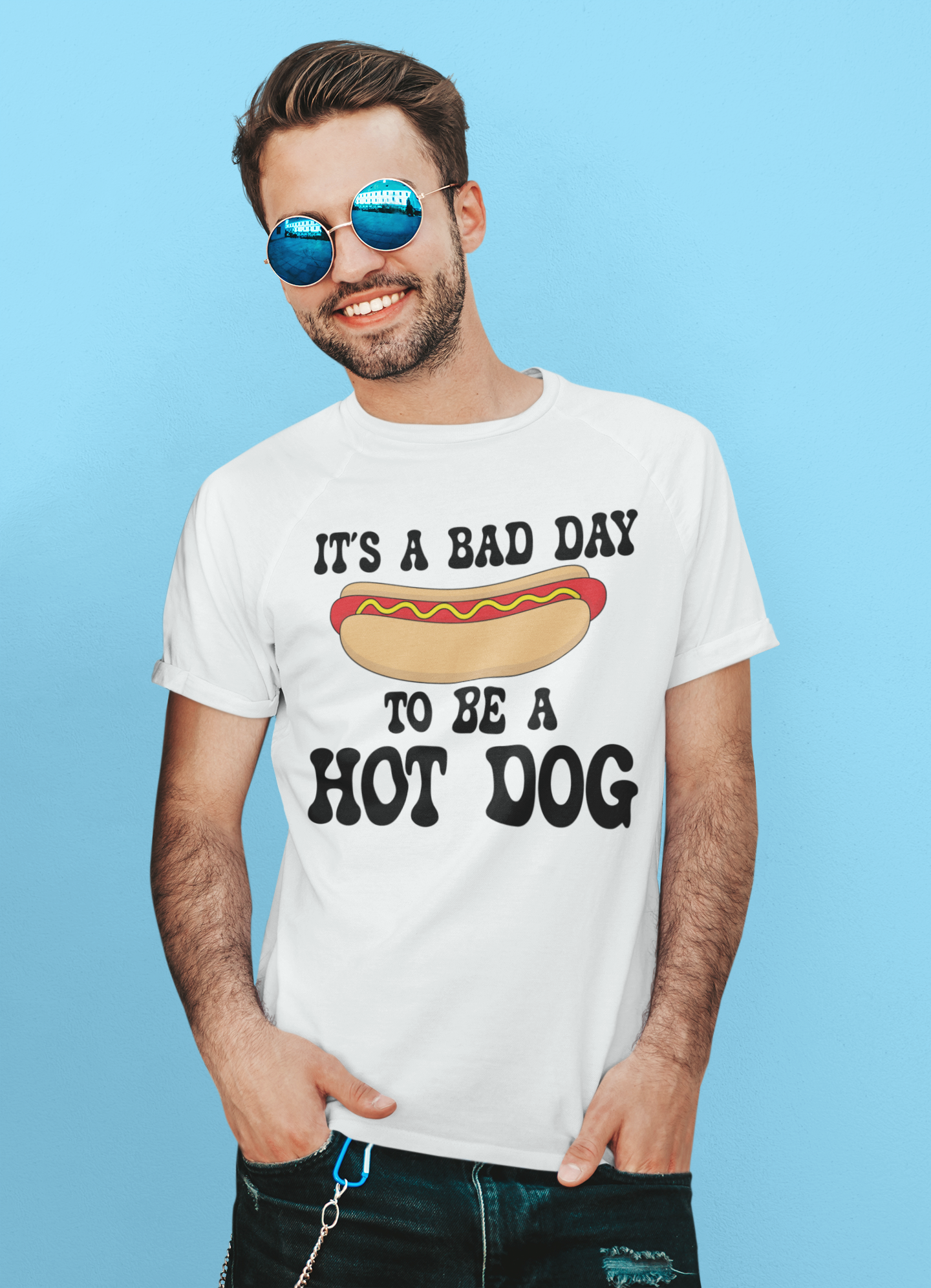A man wearing round blue mirrored sunglasses and a white graphic t-shirt that reads 'IT'S A BAD DAY TO BE A HOT DOG' with a hot dog illustration, posed against a solid light blue background.