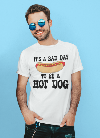 A man wearing round blue mirrored sunglasses and a white graphic t-shirt that reads 'IT'S A BAD DAY TO BE A HOT DOG' with a hot dog illustration, posed against a solid light blue background.