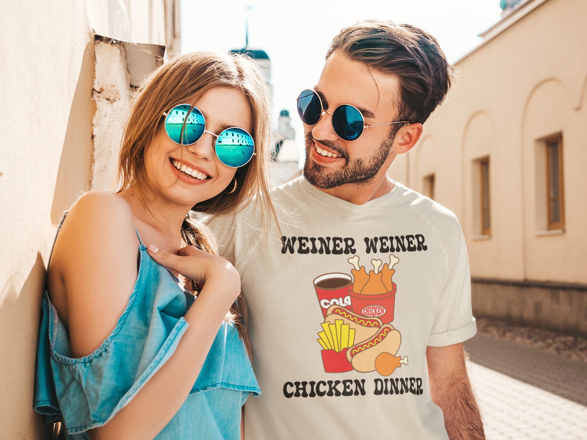 A smiling couple outdoors wearing round sunglasses. The woman wears blue mirrored sunglasses and a light blue top, while the man wears round blue mirrored sunglasses and a white graphic t-shirt that reads 'WEINER WEINER CHICKEN DINNER' with a KFC and hot dog illustration.